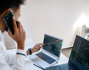 Person on a phone pointing at a laptop screen showing stock charts and market data in a bright office.
