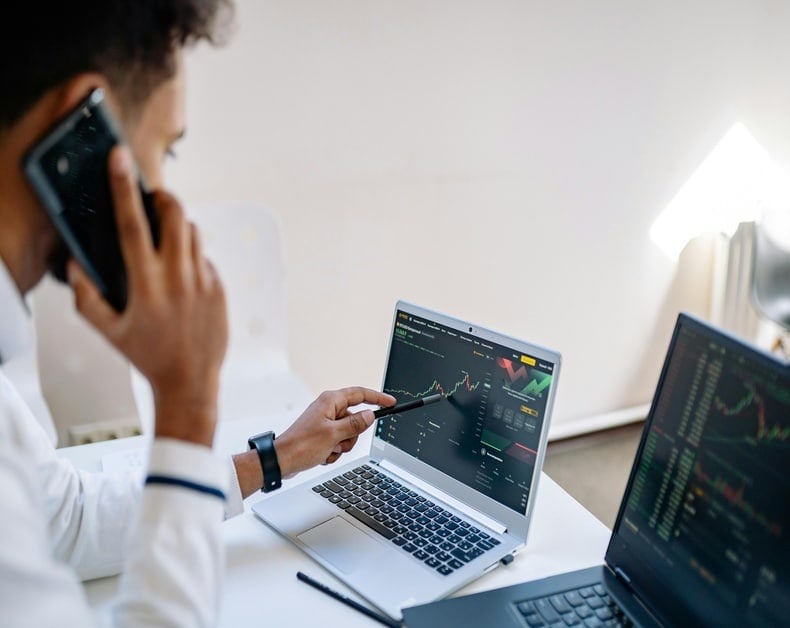 Person on a phone pointing at a laptop screen showing stock charts and market data in a bright office.