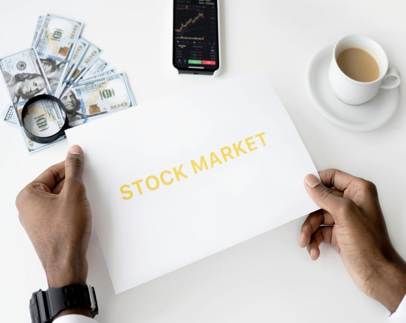 Hands hold a sheet labeled STOCK MARKET, with dollar bills, a magnifying glass, a smartphone showing stock charts, and a cup of coffee nearby.