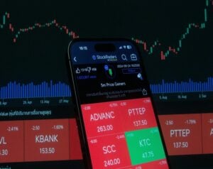 Smartphone displaying a stock trading app with red and green tiles over a stock chart background, showing prices like ADVANC and PTTEP.