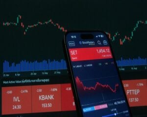 Smartphone showing a stock trading app with SET at 1,454.12 and a red price chart over a dark market dashboard.