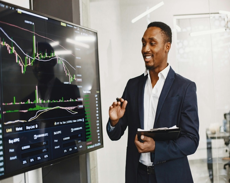 A professional man in a navy blazer presents a large screen displaying a complex stock chart with candlesticks and volume bars in an office setting, gesturing with a marker.