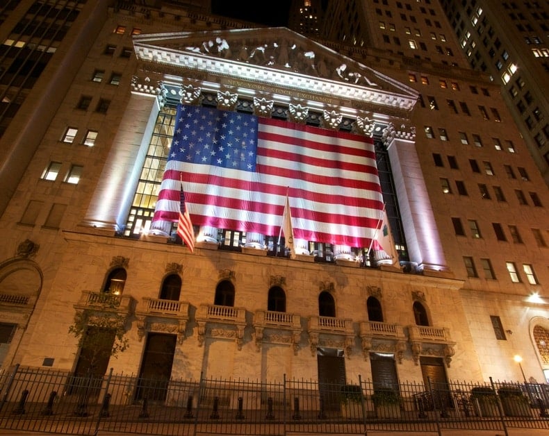 Night shot of the New York Stock Exchange facade with a large American flag draped across the front.