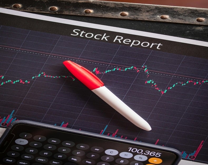 Stock market report page with a line chart, a red-and-white pen resting on it, and a smartphone showing a calculator app nearby.