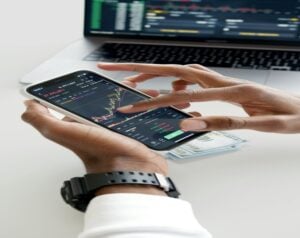 Hands holding a smartphone displaying stock charts on a trading app, with a laptop and cash on the desk in the background.
