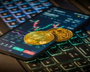 Two gold cryptocurrency coins (Bitcoin) rest on a smartphone displaying a crypto trading app with candlestick charts, placed on a backlit keyboard in low light.