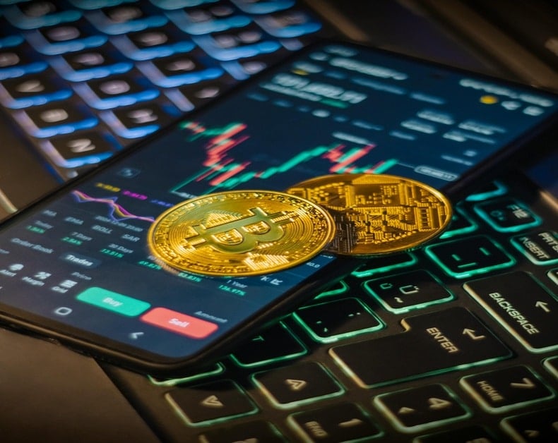 Two gold cryptocurrency coins (Bitcoin) rest on a smartphone displaying a crypto trading app with candlestick charts, placed on a backlit keyboard in low light.