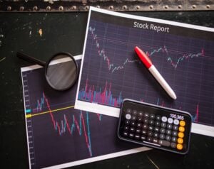 Stock analysis setup with printed charts titled 'Stock Report', a red‑white pen, magnifying glass, and a calculator on a dark desk.