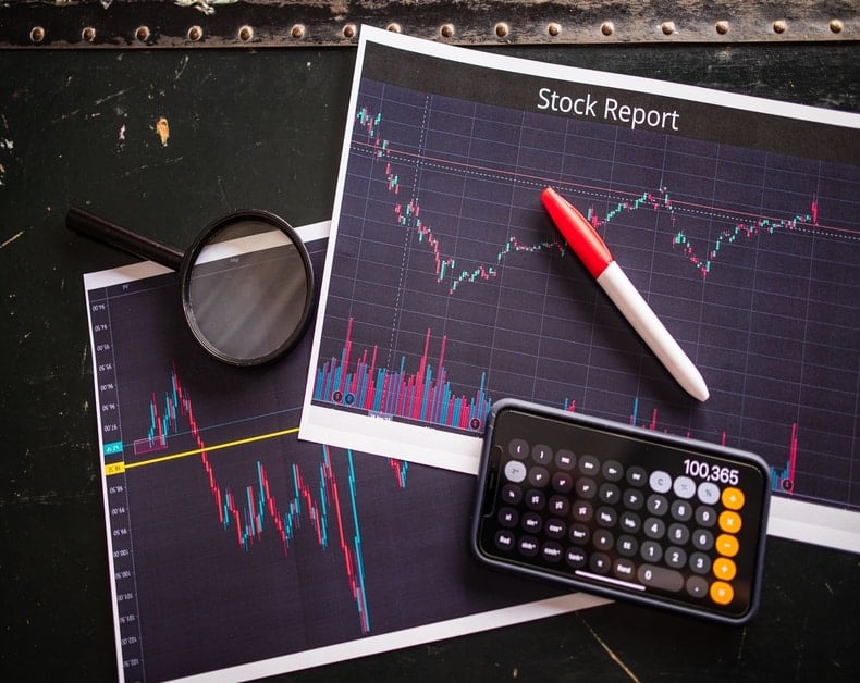Stock analysis setup with printed charts titled 'Stock Report', a red‑white pen, magnifying glass, and a calculator on a dark desk.