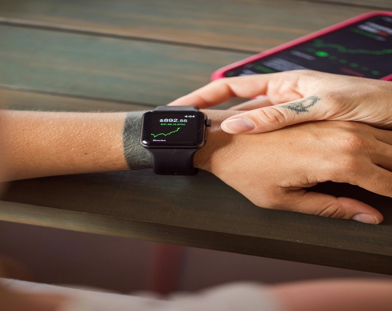 Close-up of a wrist wearing a smartwatch showing a stock price ($892.65) and a small upward graph on a wooden table surface, with a smartphone in the background.