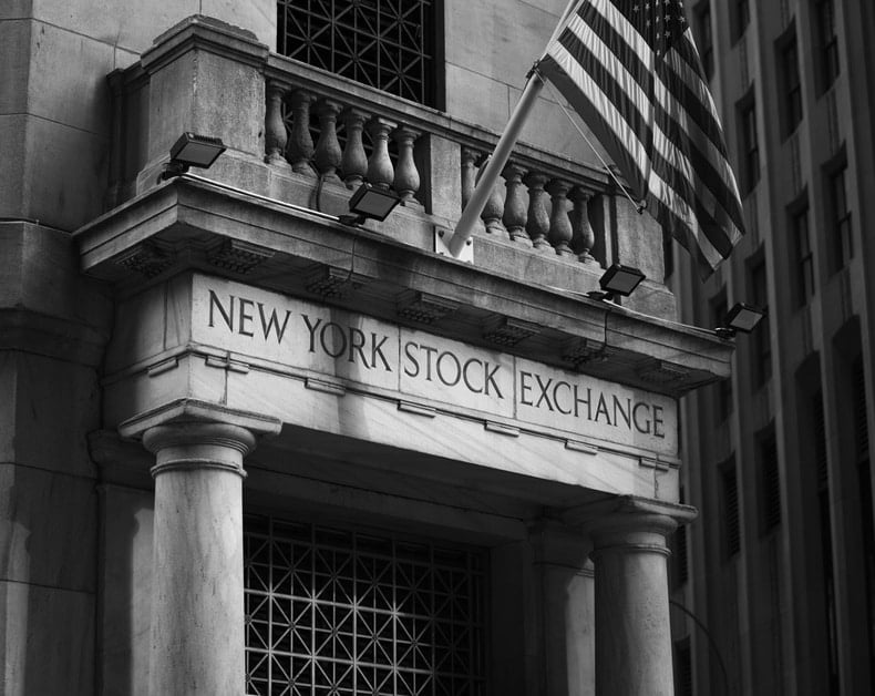 New York Stock Exchange entrance with stone columns and an American flag above it (black and white).]