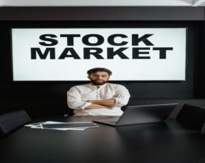 Man sits at a conference table with a laptop, arms crossed, a large 'STOCK MARKET' sign on the wall behind him.