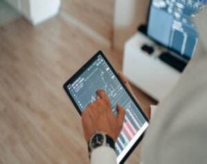 Person using a tablet to view stock/crypto trading charts in a bright room, a monitor visible in the background