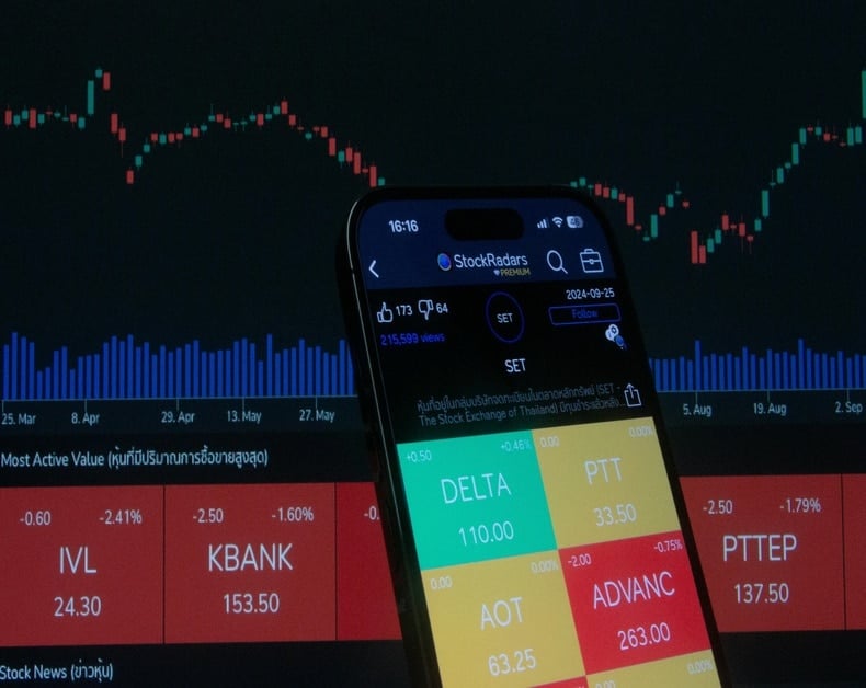 Close-up of a smartphone screen displaying a stock trading app with colorful tiles and a live market chart in the dark trading room background.