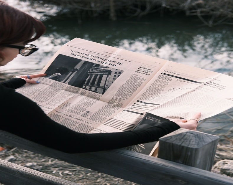 A person wearing black sleeves and glasses reads a newspaper outdoors beside a body of water.