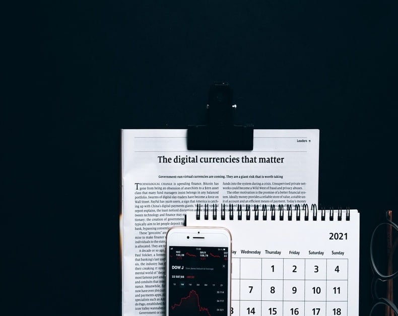 Calendar, notebook, newspaper titled 'The digital currencies that matter,' and a smartphone showing a market chart on a dark desk backdrop.
