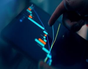 Finger points at a touchscreen showing a colorful stock candlestick chart on a tablet screen.