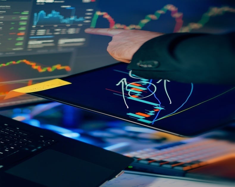 Businessperson's hand points at a tablet showing colorful stock charts, with monitors and keyboard in the background.