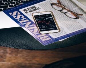 Newspaper spread with 'The Future of Business' headline, a smartphone showing crypto charts, and a pair of glasses on the page.