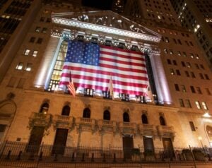 Nighttime view of the New York Stock Exchange building with a giant American flag draped across its façade, flanked by two smaller flags.