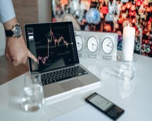 Person points at a laptop screen displaying a stock chart on a white desk with clocks, a candle, and a smartphone nearby.