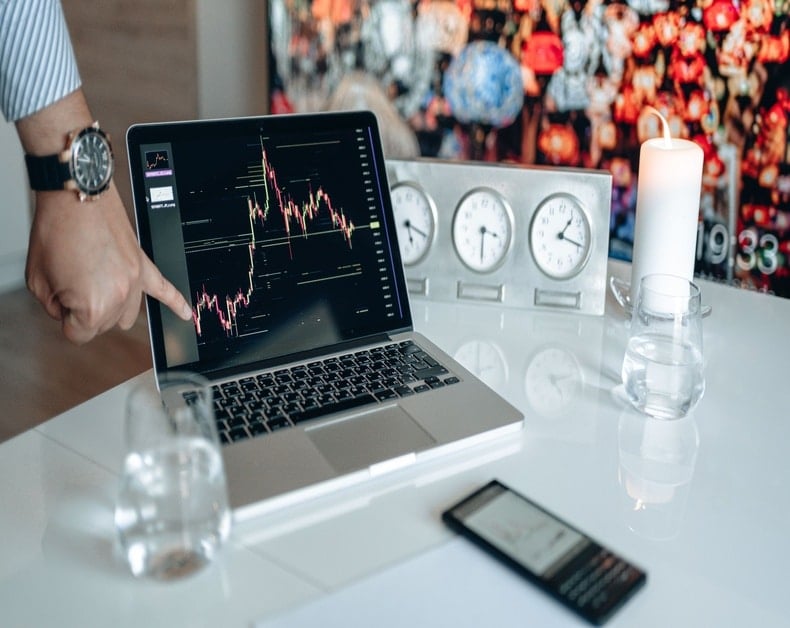 Person points at a laptop screen displaying a stock chart on a white desk with clocks, a candle, and a smartphone nearby.