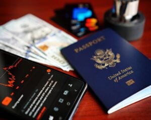 Blue US passport, cash, credit cards, and a smartphone displaying a financial app on a wooden desk base.