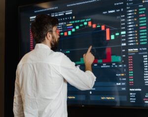 Man with a beard in a white shirt points at a large screen displaying candlestick stock charts and volume bars on a trading dashboard.