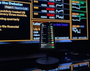 Smartphone on a stand displaying a stock market app with green and red numbers, set against a large illuminated financial ticker board in the background.