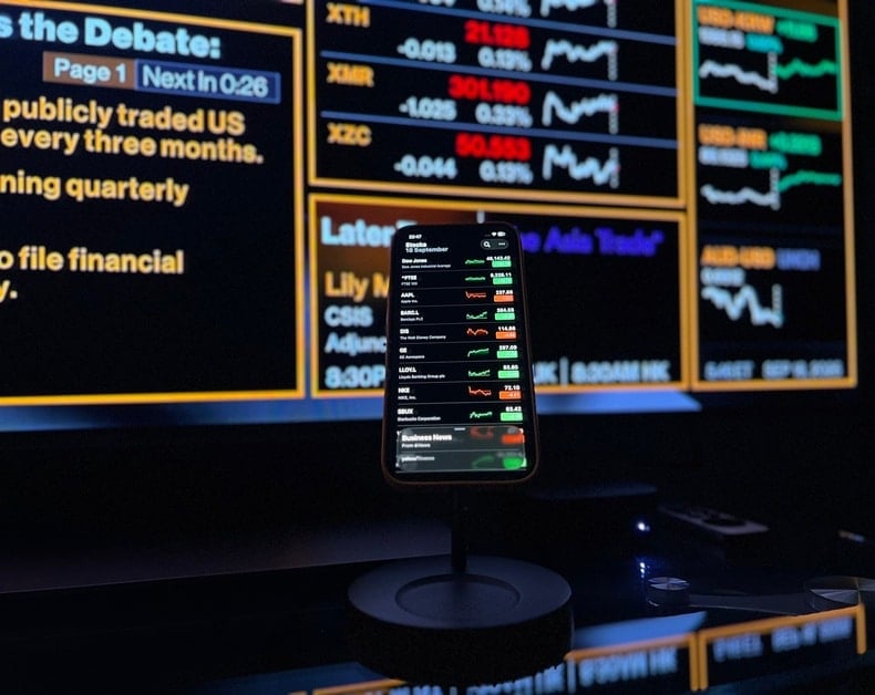 Smartphone on a stand displaying a stock market app with green and red numbers, set against a large illuminated financial ticker board in the background.