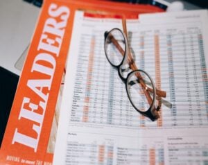 Open financial report with stock tables and a pair of round glasses resting on the pages, beside an orange magazine titled TRADERS/HEADERS.