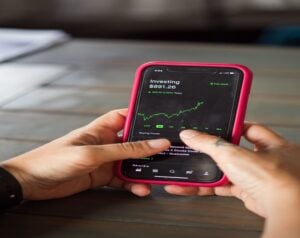 Person holding a smartphone in a pink case showing an investing app with a green stock chart and a balance of $891.26 on screen.