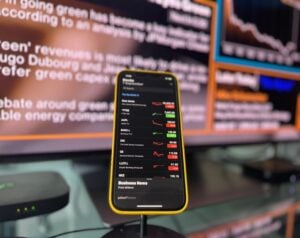 Smartphone with stock market app displayed in focus, showing tickers and red/green arrows, on a desk with blurred monitors in the background