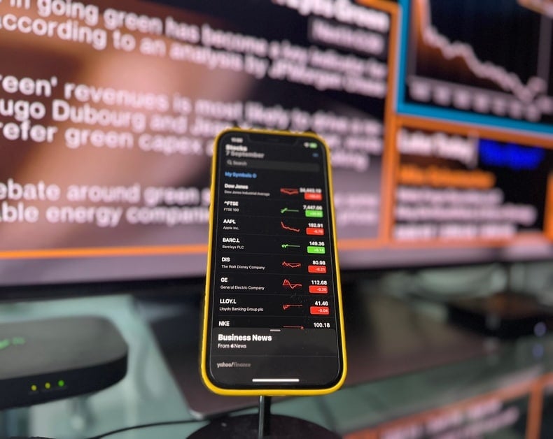 Smartphone with stock market app displayed in focus, showing tickers and red/green arrows, on a desk with blurred monitors in the background
