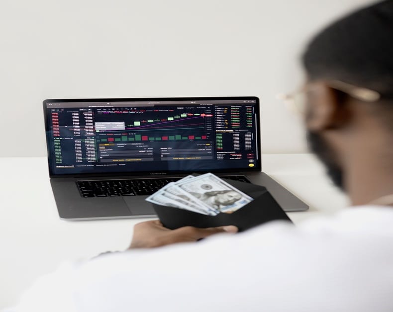 Person with a wallet holding cash in front of a laptop displaying a stock trading dashboard.