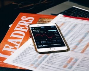 Smartphone showing a Bitcoin price chart placed on top of financial documents and a finance magazine on a desk.