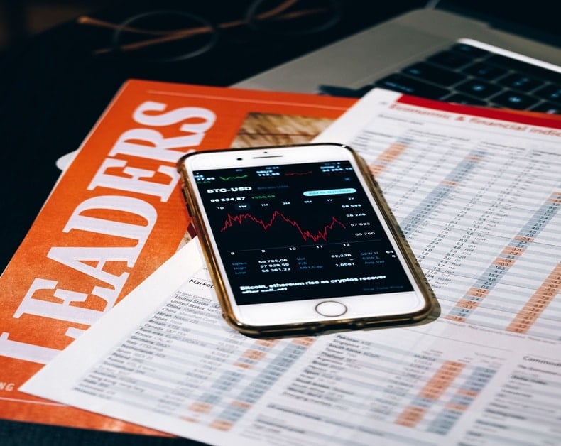 Smartphone showing a Bitcoin price chart placed on top of financial documents and a finance magazine on a desk.
