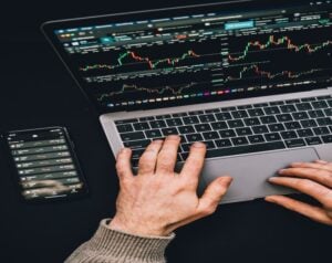 Close-up of hands on a laptop displaying stock charts, with a smartphone showing a trading app beside it.