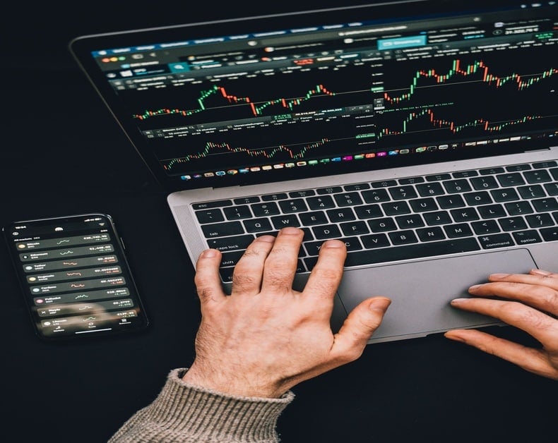 Close-up of hands on a laptop displaying stock charts, with a smartphone showing a trading app beside it.