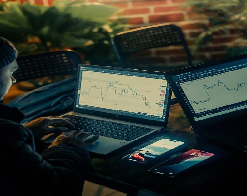 Person working on two laptops displaying stock charts, with a smartphone nearby.