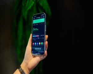 Person holding a smartphone showing a stock trading app and chart on screen against a dark background.