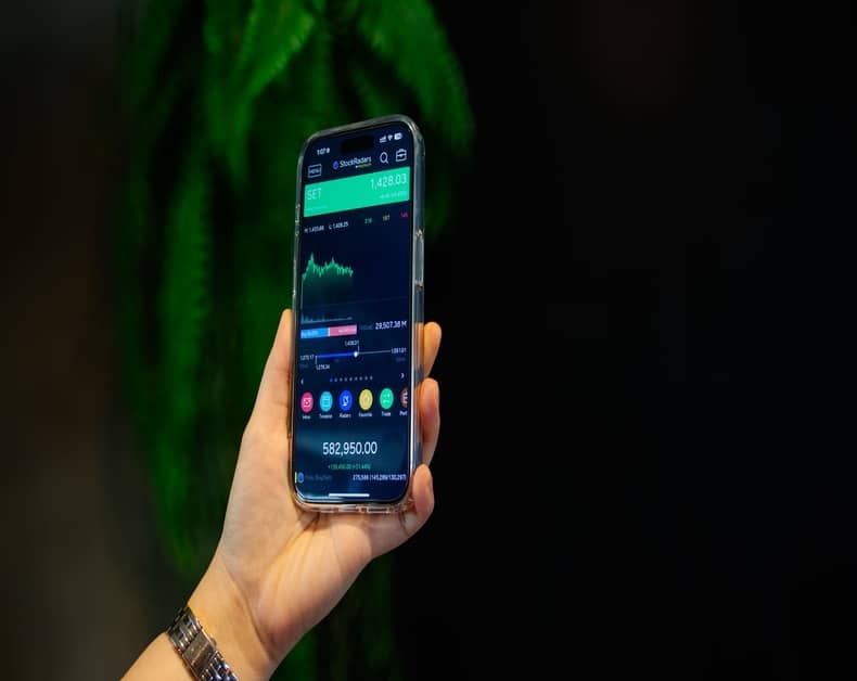 Person holding a smartphone showing a stock trading app and chart on screen against a dark background.