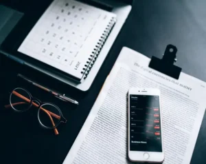 A desk scene with a spiral notebook, glasses, a pen, a calendar, and a smartphone displaying stock data over an open article on a clipboard.