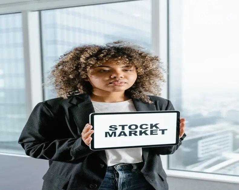Woman in a blazer holds a tablet reading 'STOCK MARKET' in a bright office setting, presenting financial information