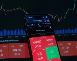 Smartphone displaying a stock trading app with red/green tiles (ADVANC 263, PTTEP 137.5, KTC 47.75) in front of a blue/green market chart background.