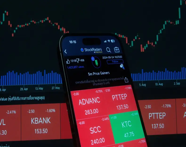 Smartphone displaying a stock trading app with red/green tiles (ADVANC 263, PTTEP 137.5, KTC 47.75) in front of a blue/green market chart background.