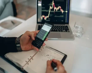 Person using a calculator while reviewing a notebook with stock charts on a laptop screen.