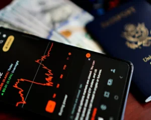 Close-up of a smartphone showing a red line stock chart on a dark screen, with blurred money and a passport in the background.