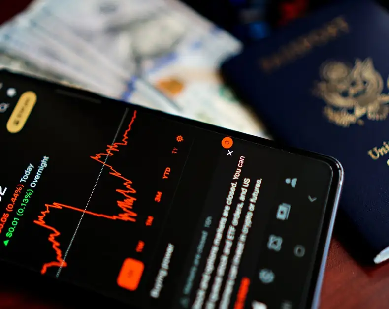 Close-up of a smartphone showing a red line stock chart on a dark screen, with blurred money and a passport in the background.