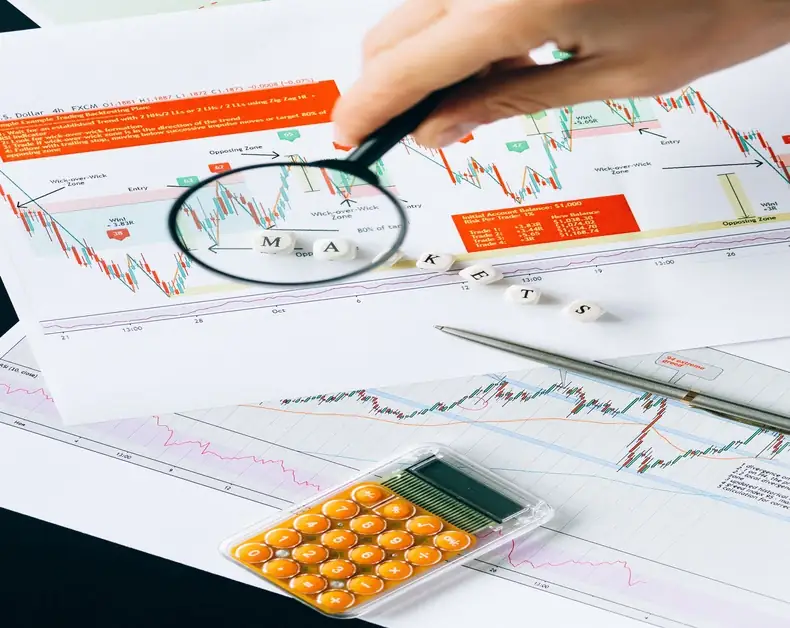 Person analyzes stock charts with a magnifying glass over printed financial data and graphs on a desk.